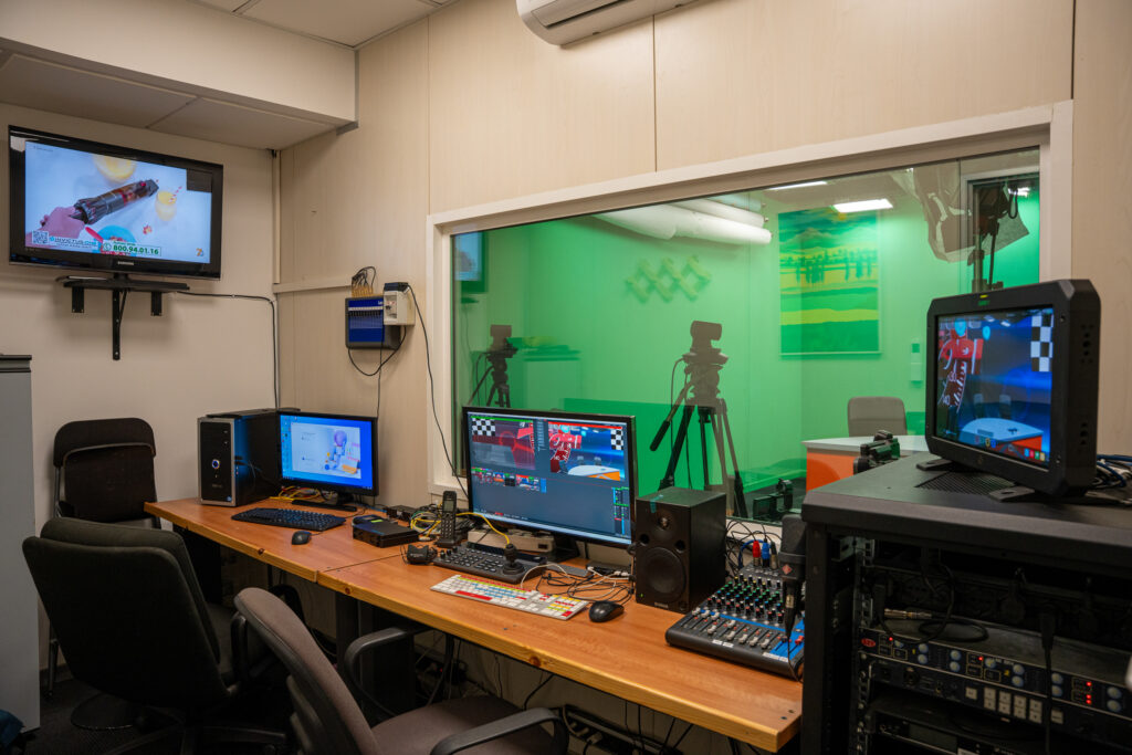 Regia dello studio TV con vetrata sul set green screen, monitor e mixer audio